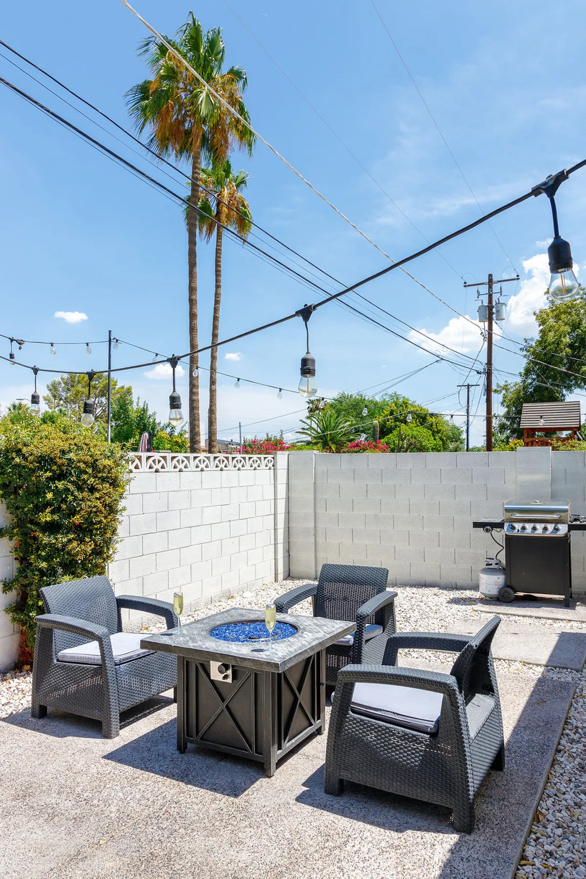 An inviting outdoor seating area centered around a modern fire pit with blue glass, flanked by lounge seating, a grill station, palm trees, and string lighting. The setup creates a cozy spot for evening gatherings and s’mores under the stars.