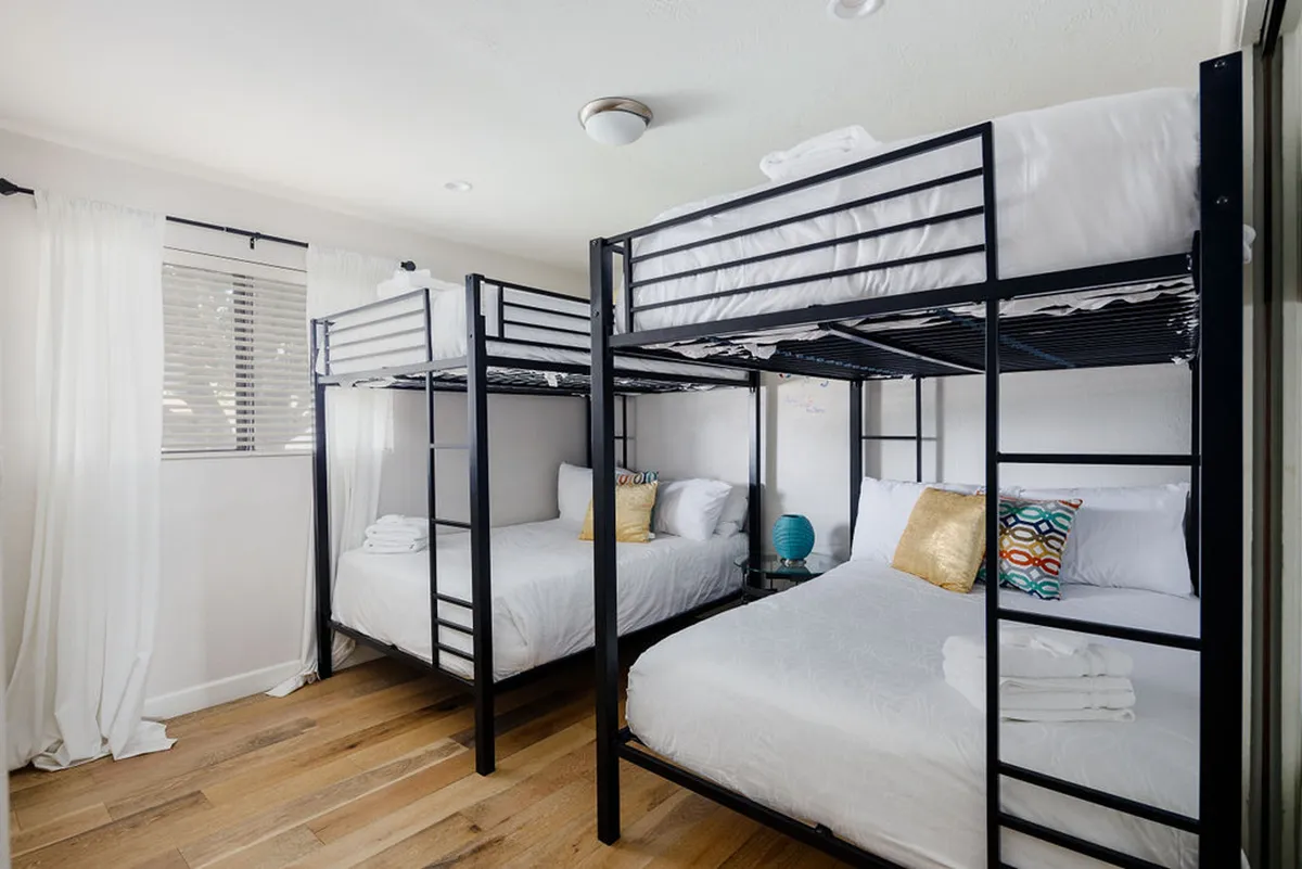 A cozy and playful bunk room complete with bunk beds, decorative pillows, and a nightstand with a lamp. Sunlight streams in, setting the scene for fun sleepovers, late-night reading, and relaxed mornings—making it perfect for kids or groups.