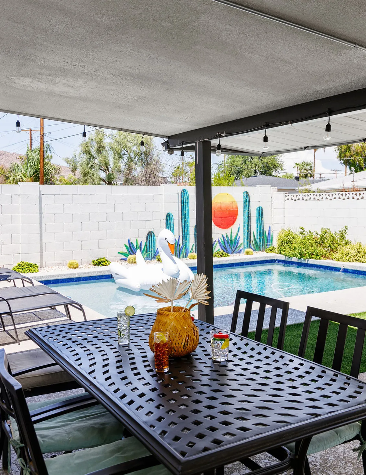 An inviting outdoor dining table is set beside a sparkling swimming pool, with a colorful wall mural and pool float adding a playful, social vibe. The setting is perfect for sharing meals or relaxing poolside with friends during the day or at sunset.