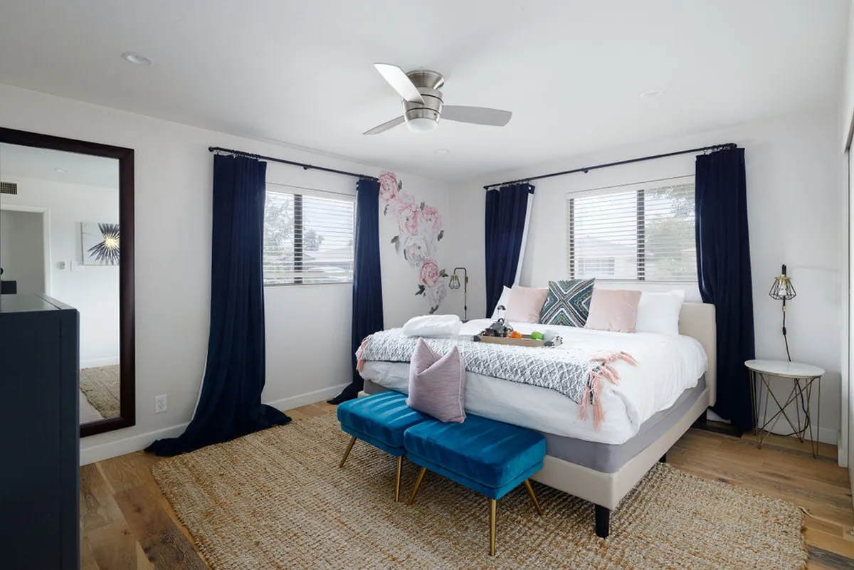 A serene and stylish primary bedroom with a king-sized bed, floral wall art above the headboard, two blue ottomans at the foot of the bed, and a large mirror that amplifies the natural light streaming into the room. The space feels restful and inviting, ideal for those seeking a luxurious retreat.