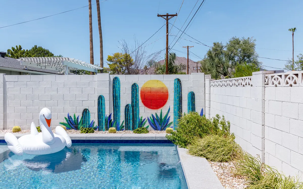 A vibrant backyard scene featuring a sparkling swimming pool, an inflatable swan, a colorful mural with sun and cacti, and lush landscaping. The playful setting is perfect for sunbathing, swimming, and enjoying outdoor relaxation with friends.