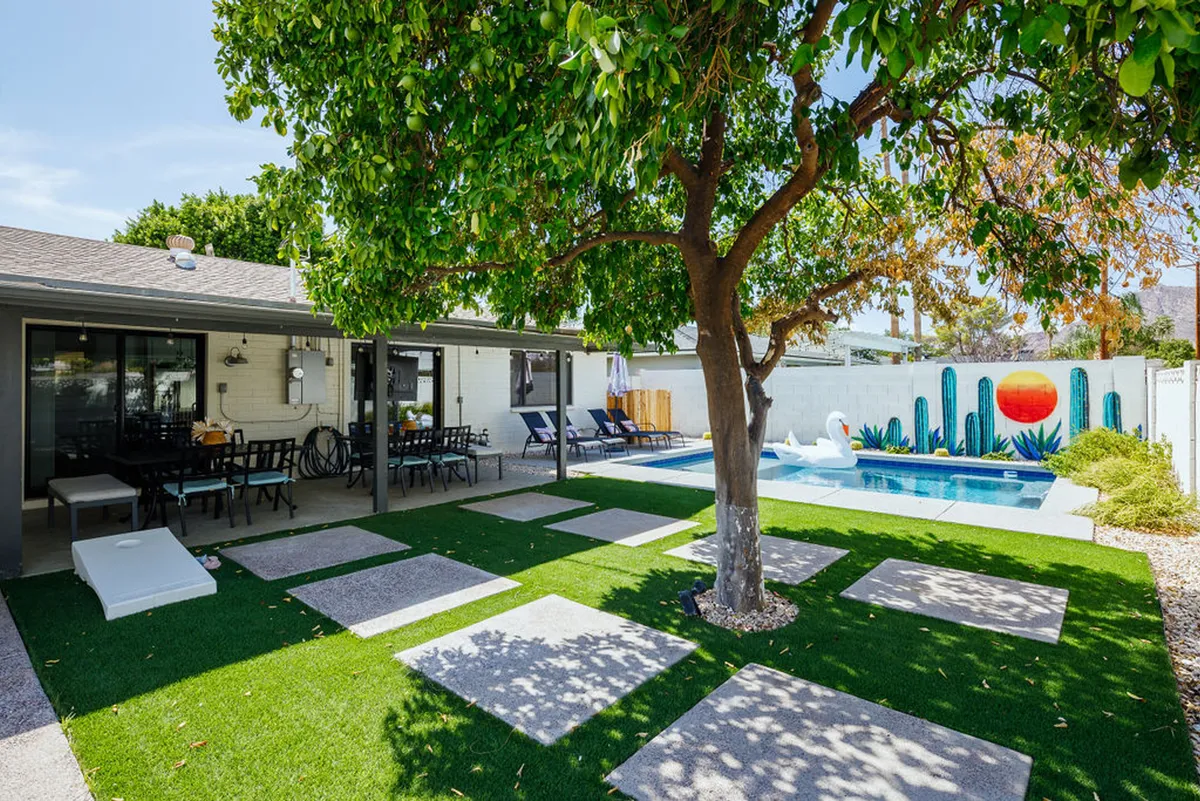 A cheerful backyard scene with a swimming pool, colorful mural featuring sun and cacti, an outdoor dining area, lush green lawn, and a large shade tree. The space is set up for swimming, sunbathing, dining, and relaxing—ideal for families to gather after a day of exploring.