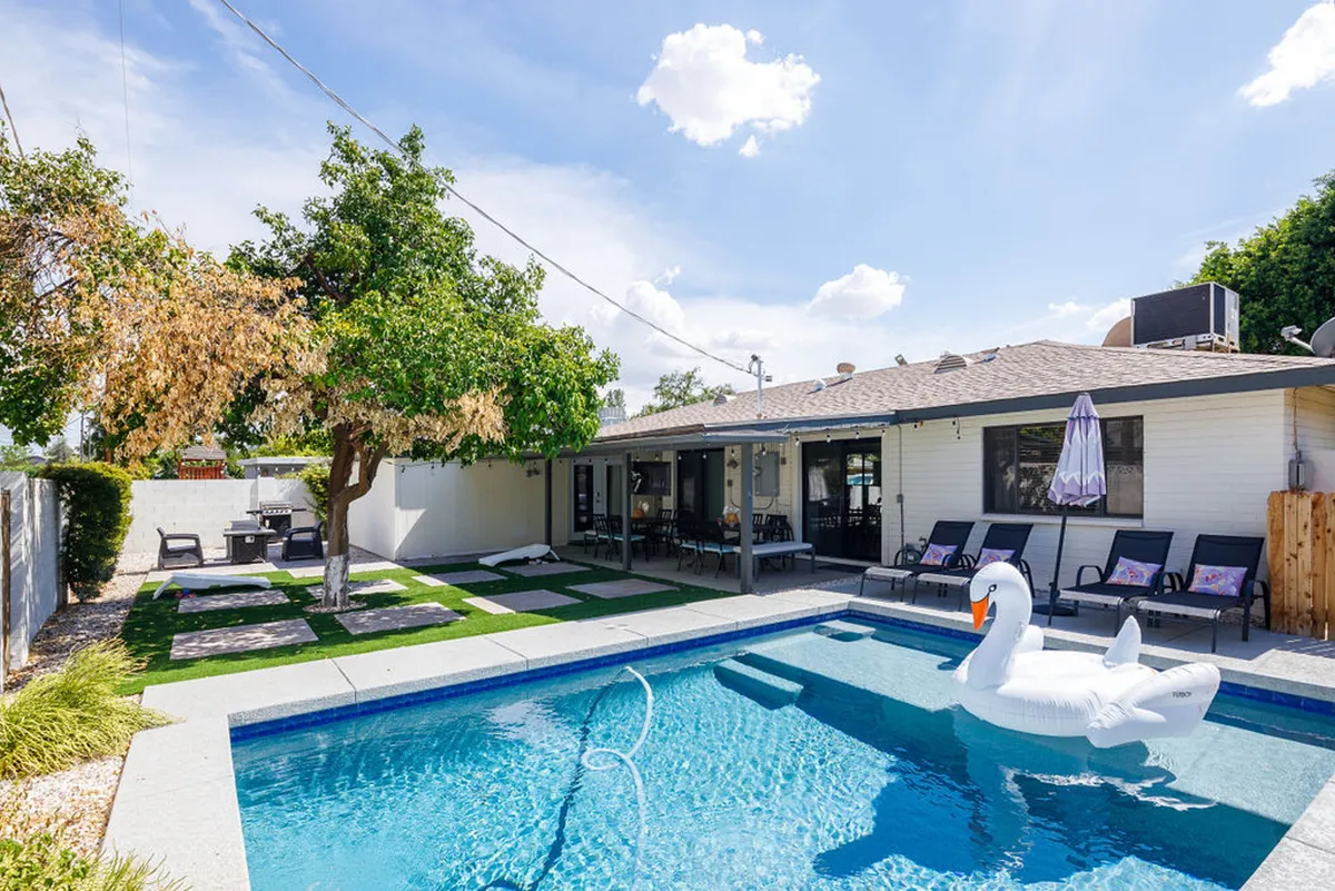 A cheerful backyard view with a sparkling pool, floating swan, lounge chairs shaded by a leafy tree, and an adjacent outdoor dining area. The scene radiates fun and leisure, with plenty of space for families and friends to gather and play.