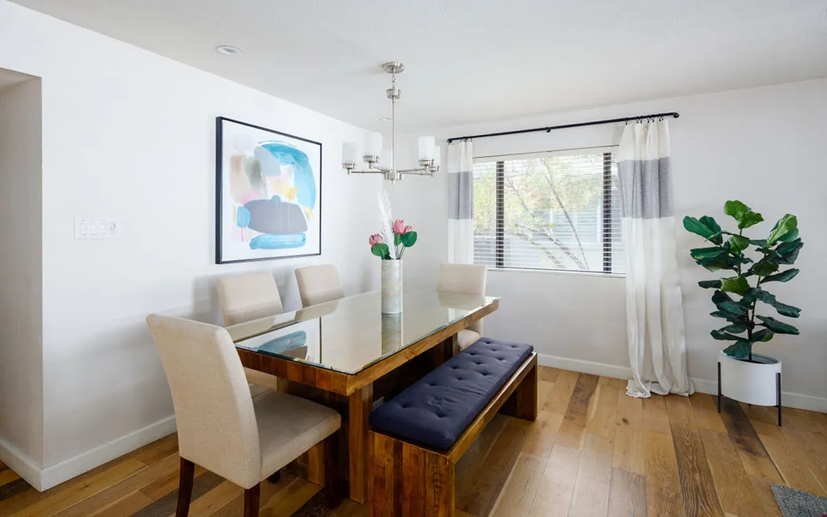 A stylish dining area featuring a glass-top table set for a meal, statement artwork on the wall, a potted plant for warmth, and an elegant chandelier overhead. The setting is inviting and versatile, perfect for shared meals, game nights, or planning adventures as a group.