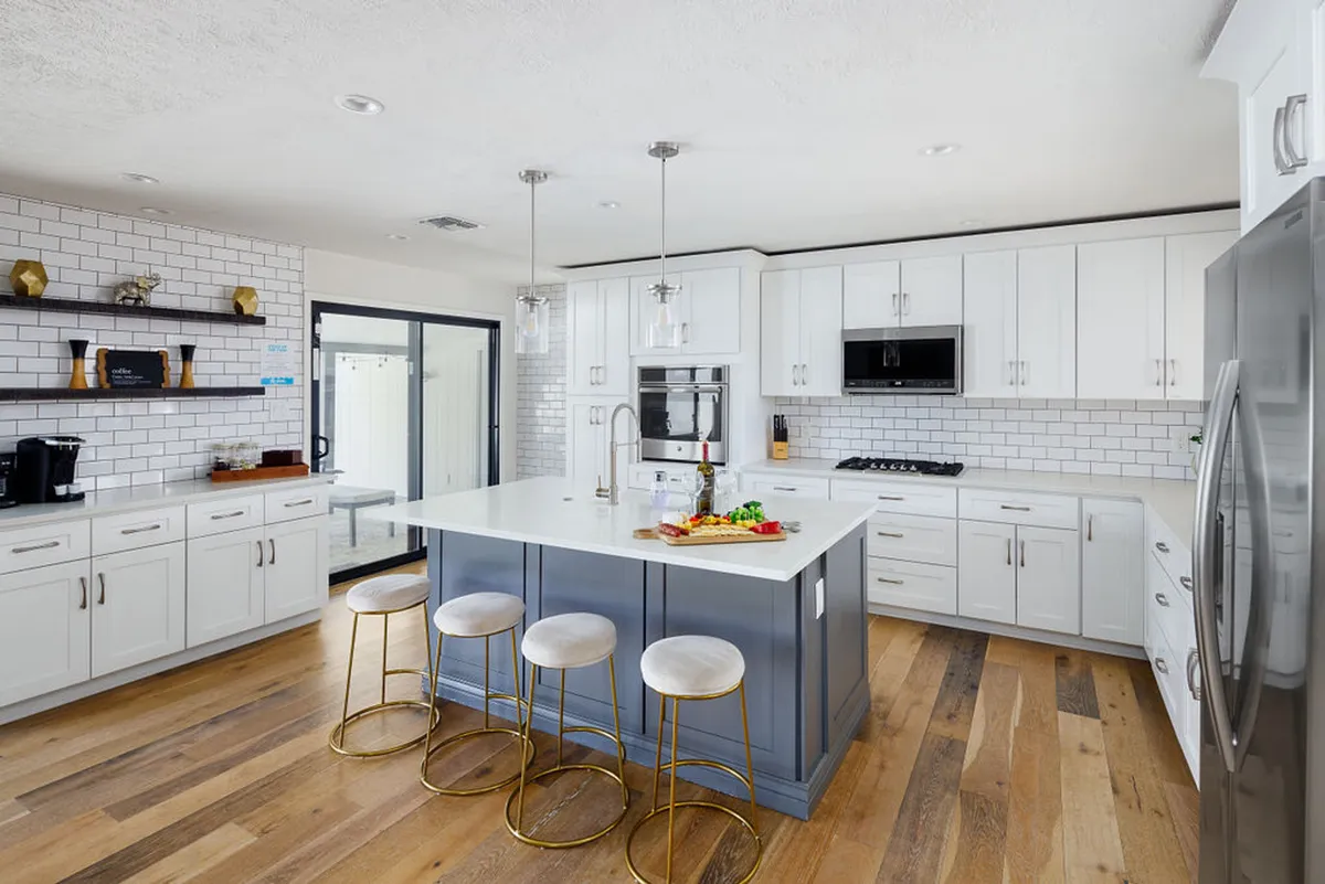 A kitchen scene focused on a central island with a cutting board and fresh vegetables, surrounded by open shelving with decorative items and modern stainless appliances. The layout hints at an inviting, functional space where meal prep and group participation are encouraged.